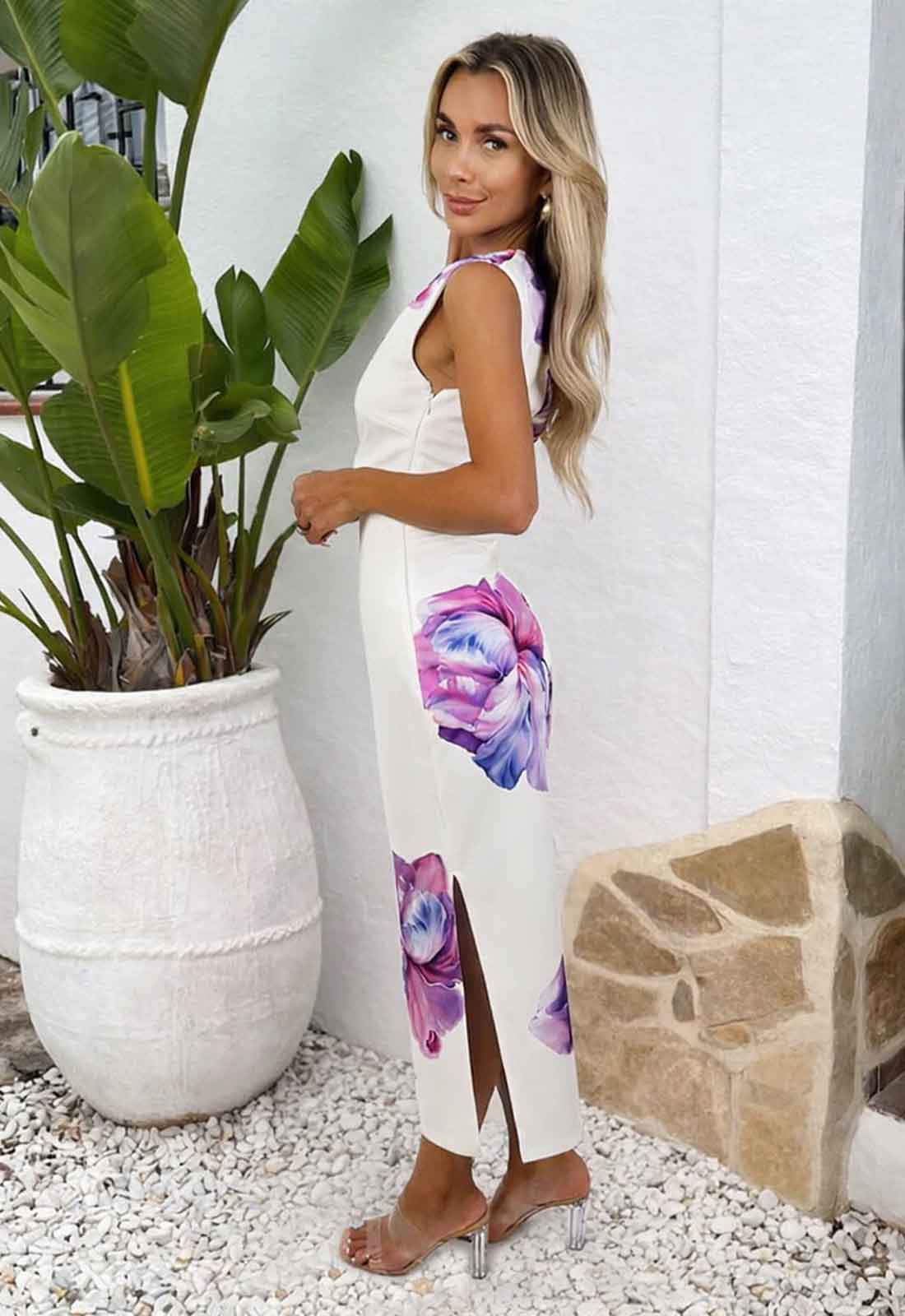 Woman in a white dress with floral patterns standing next to a potted plant.