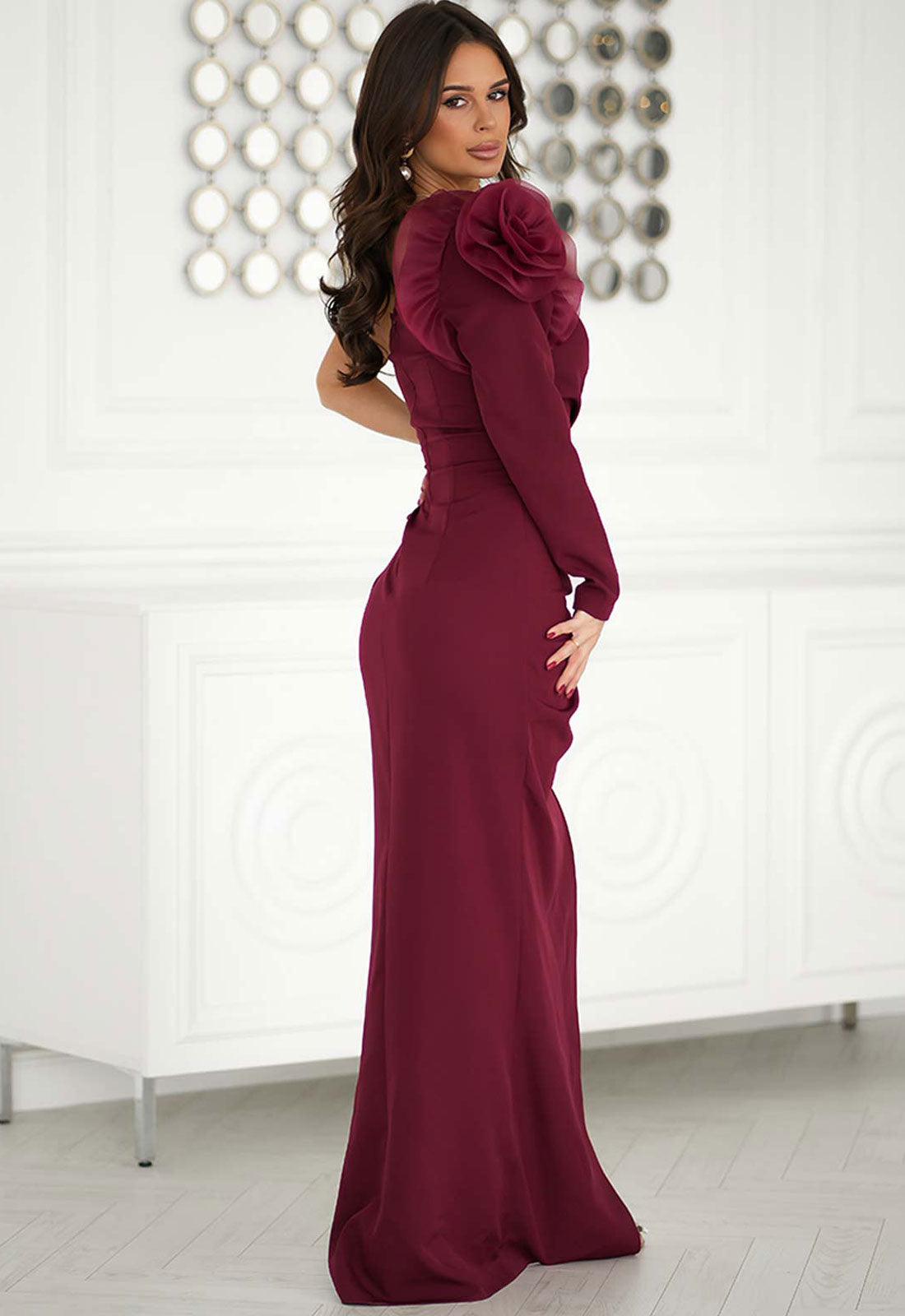 Woman wearing a burgundy evening gown with ruffled sleeves against a white wall.