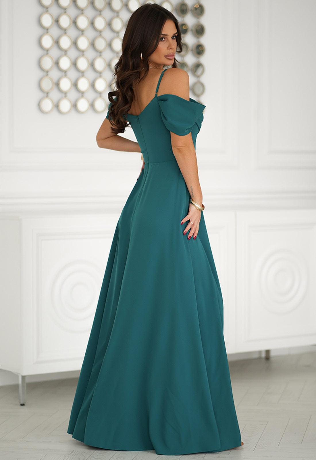 Woman wearing a teal off-shoulder gown in a room with decorative wall.
