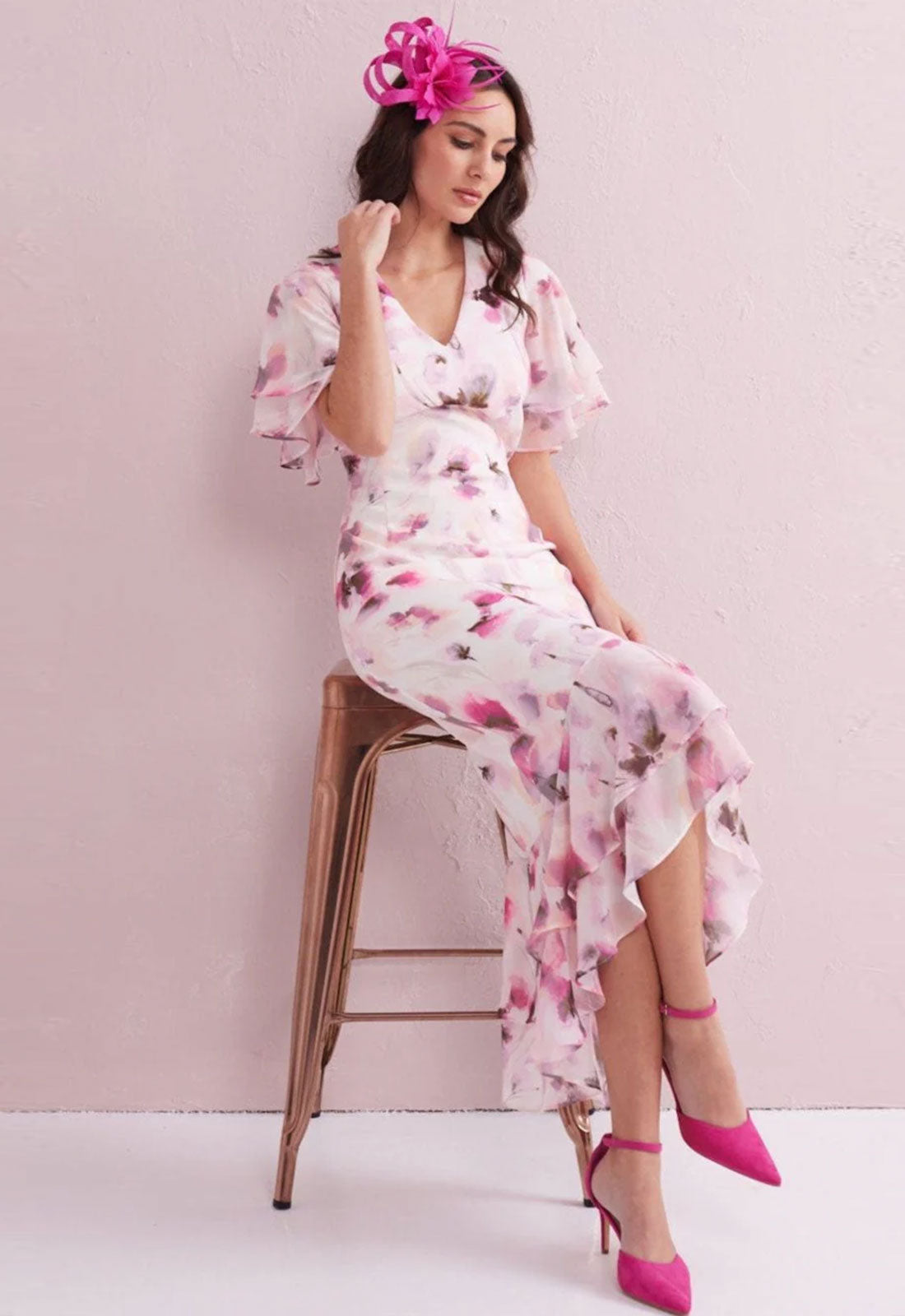 Woman in a floral dress sitting on a stool against a pink background
