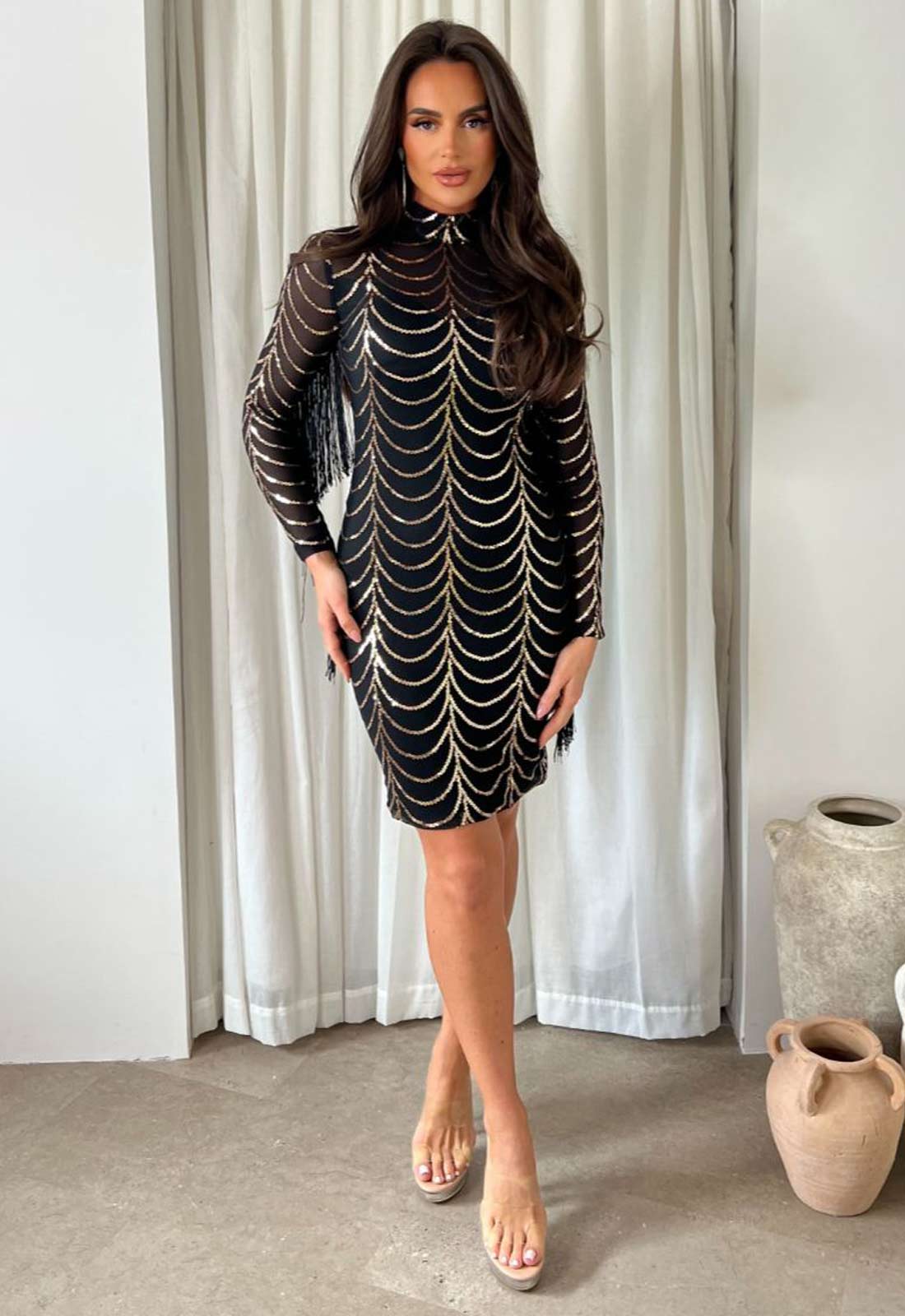 Woman wearing a black dress with white patterns in a room with curtains and vases.