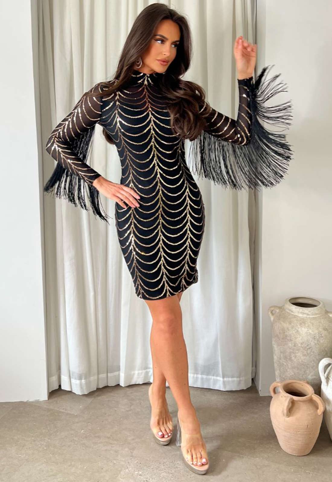Woman wearing a black dress with fringe details in a room with white curtains and decorative vases.