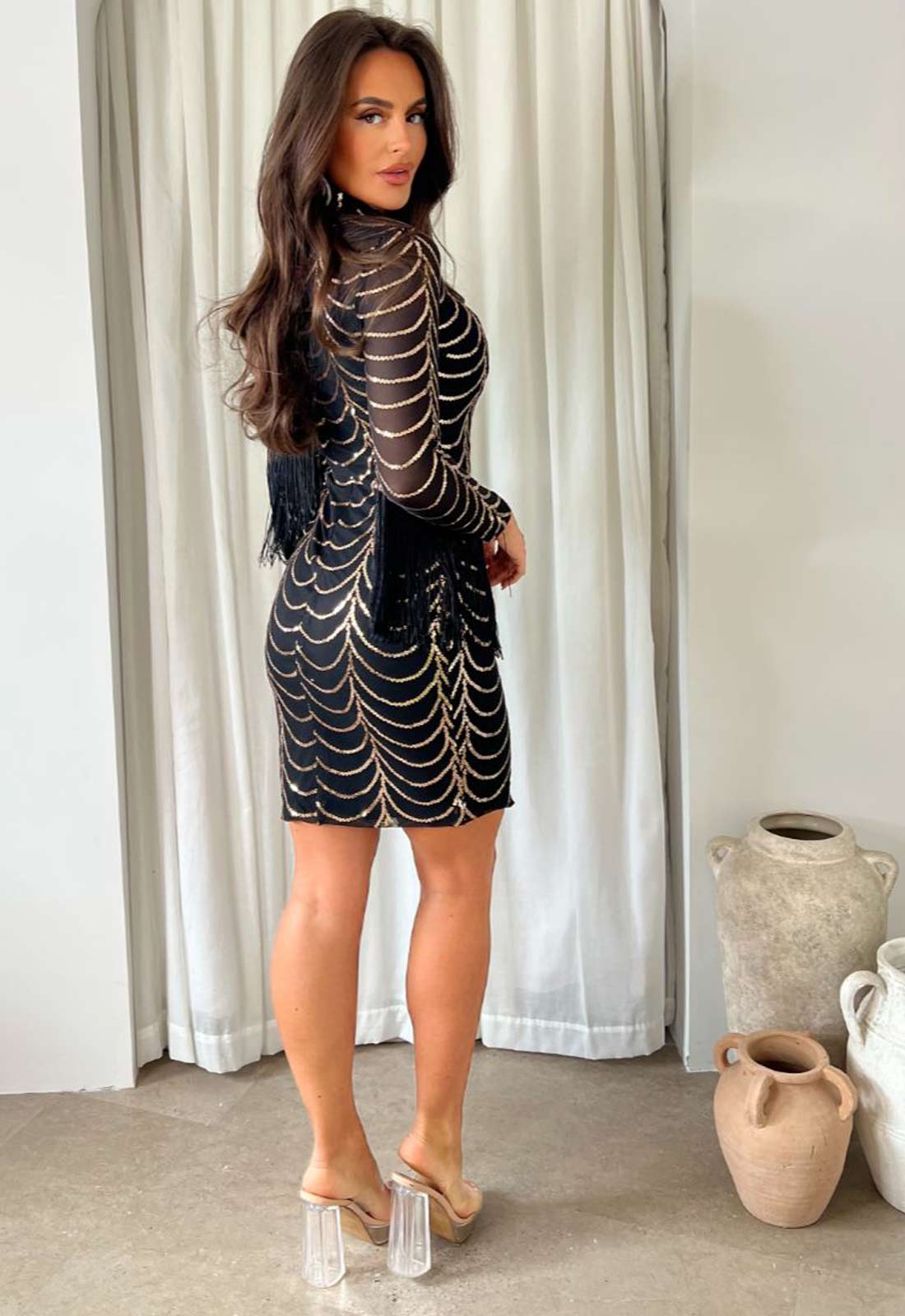 Woman wearing a black dress with gold patterns in a room with white curtains and decorative vases.