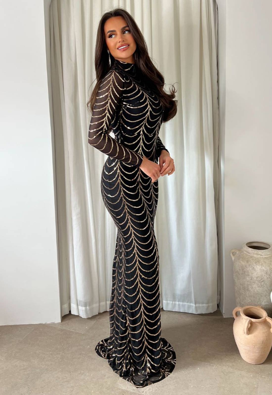 Woman in a black and gold patterned dress standing in front of white curtains.