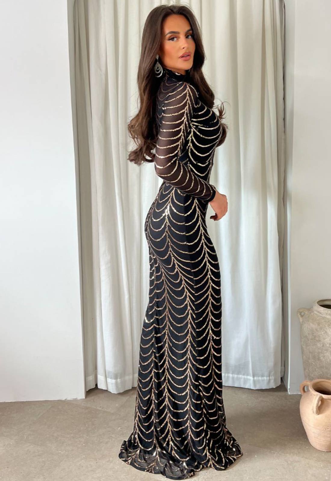 Woman wearing a black evening gown with gold detailing in a room with white curtains.