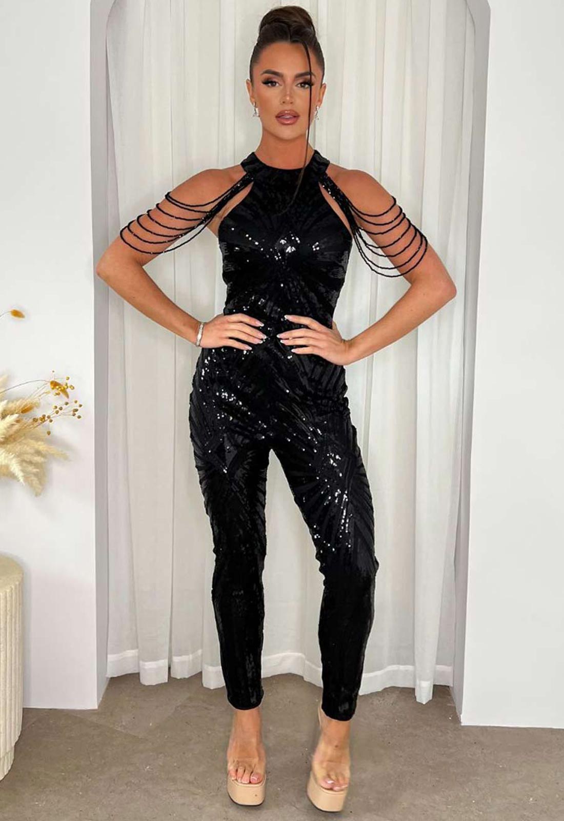 Woman wearing a black sequin jumpsuit with sheer sleeves against a white curtain background