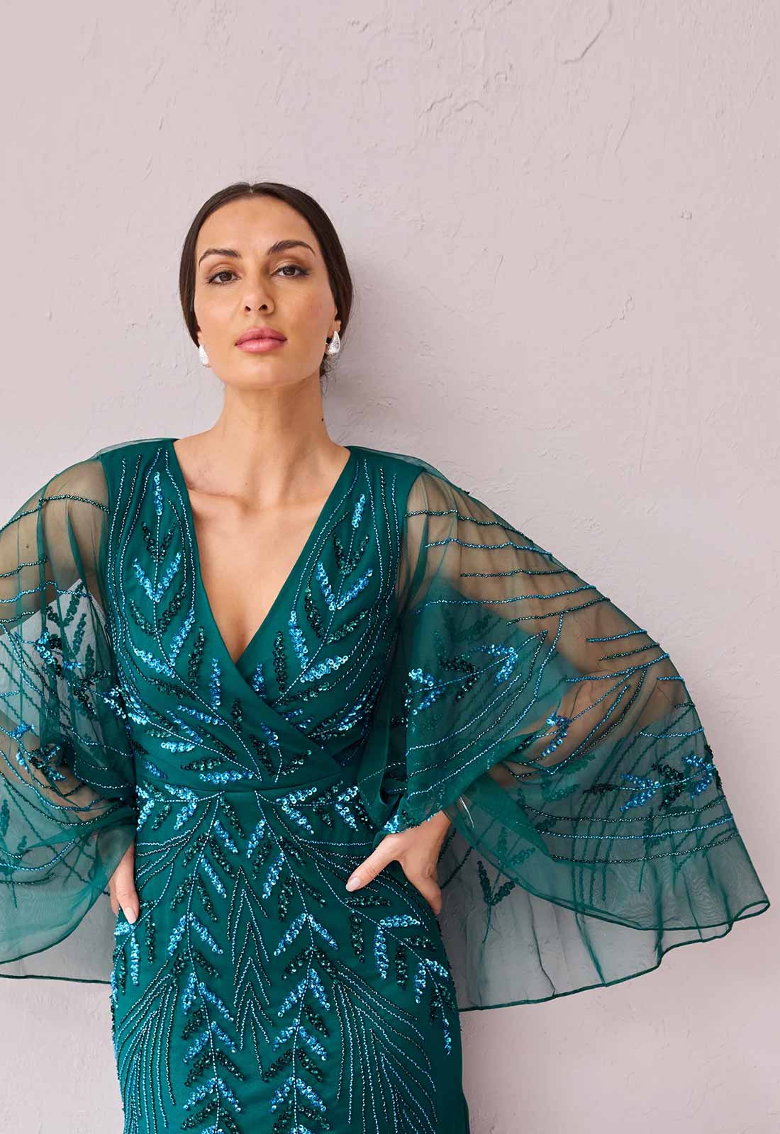 A woman wearing a teal green sequined evening gown with sheer bell sleeves and geometric patterns, posing against a textured light pink wall.