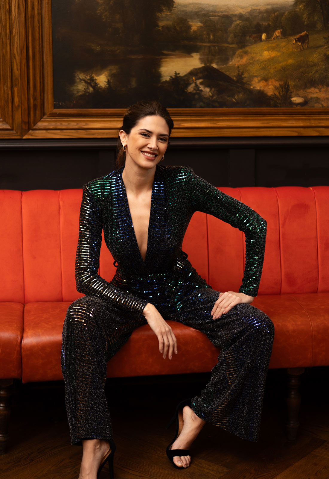 Woman in a black sequin outfit sitting on a red couch with a painting in the background