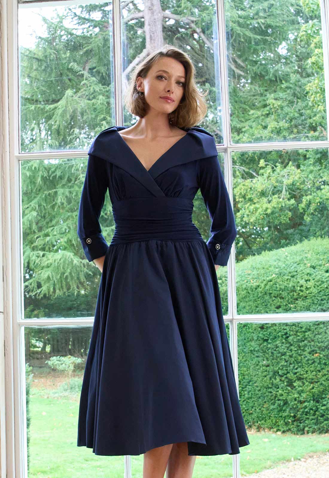 Woman wearing a navy blue dress with a white headband in front of a window with greenery outside.
