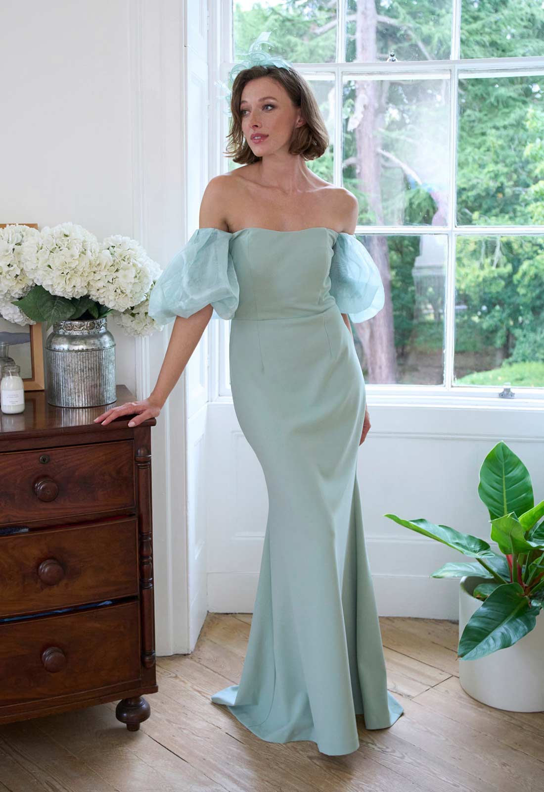 Woman in a light green off-shoulder dress standing in a room with wooden furniture and plants.