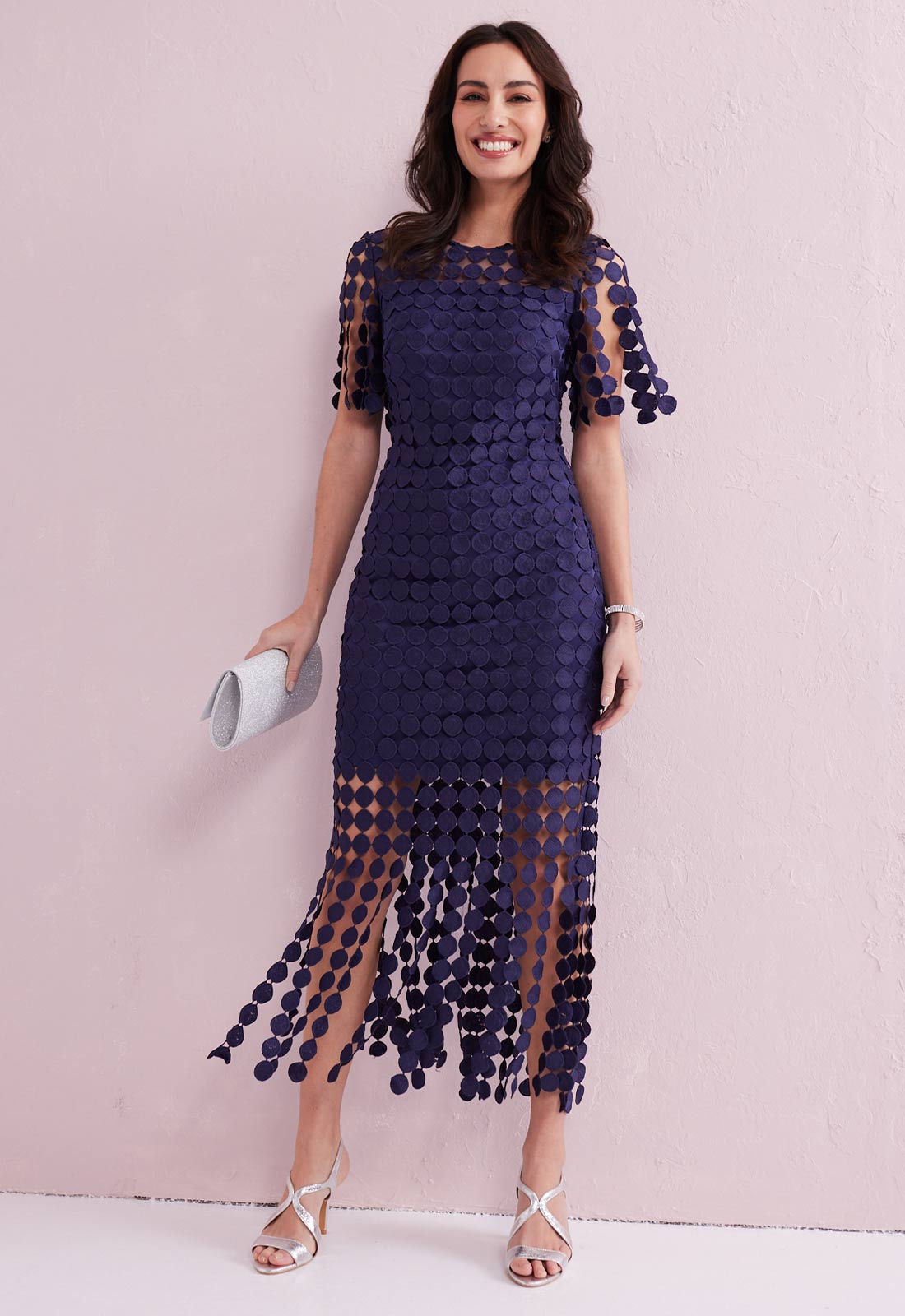 Woman wearing a navy blue dress with sheer sleeves and hem against a pink background