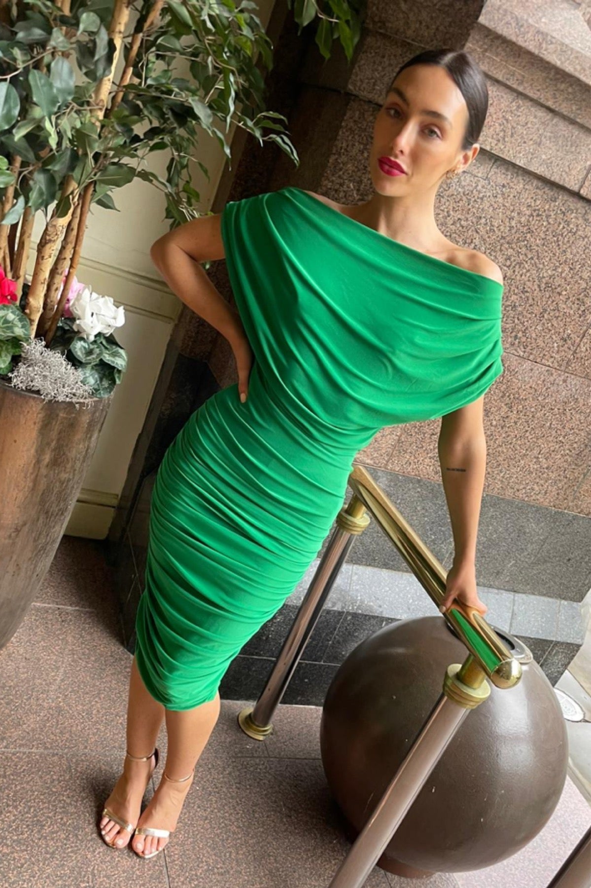 Woman in a green dress standing next to a plant and decorative object indoors.