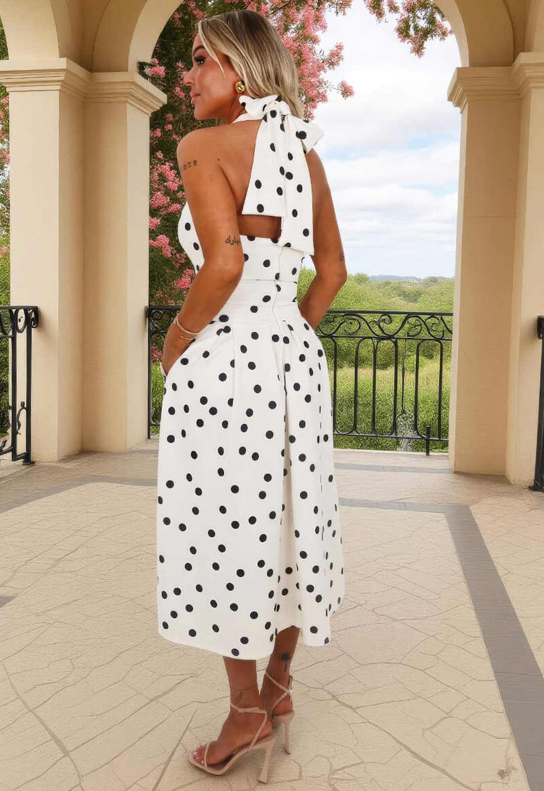 Woman in a white polka dot dress standing in an outdoor setting with columns and greenery.