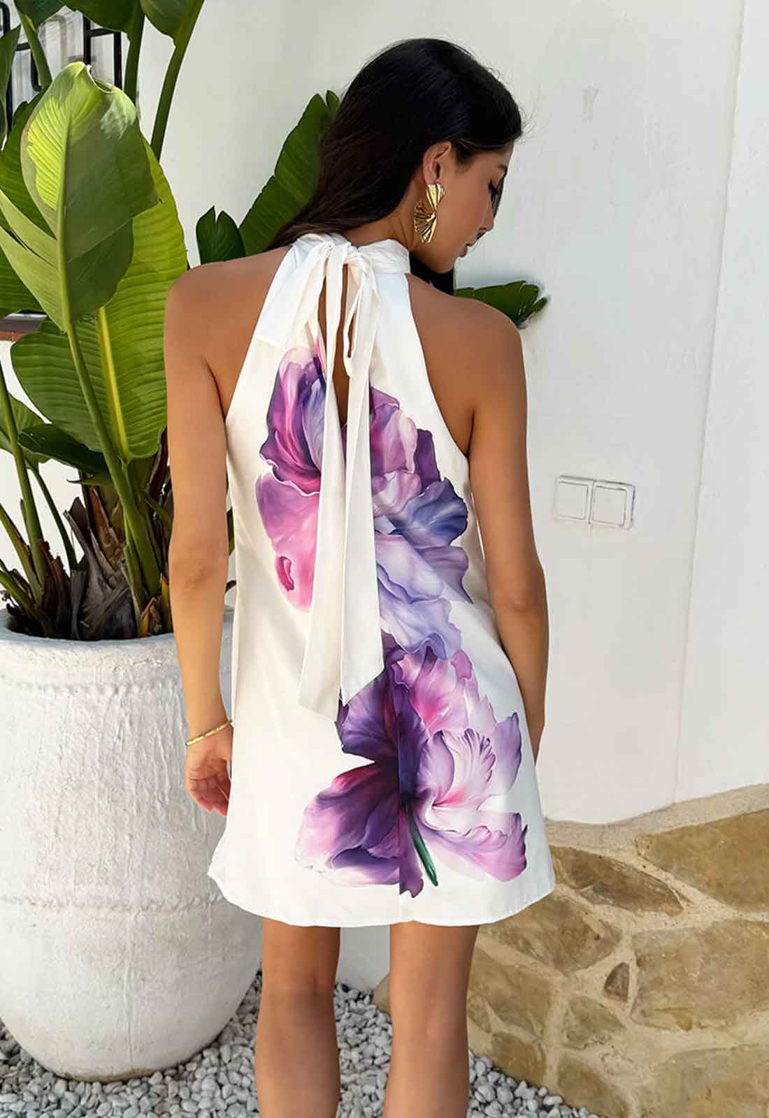 Woman wearing a white dress with purple floral design, standing outdoors near plants.