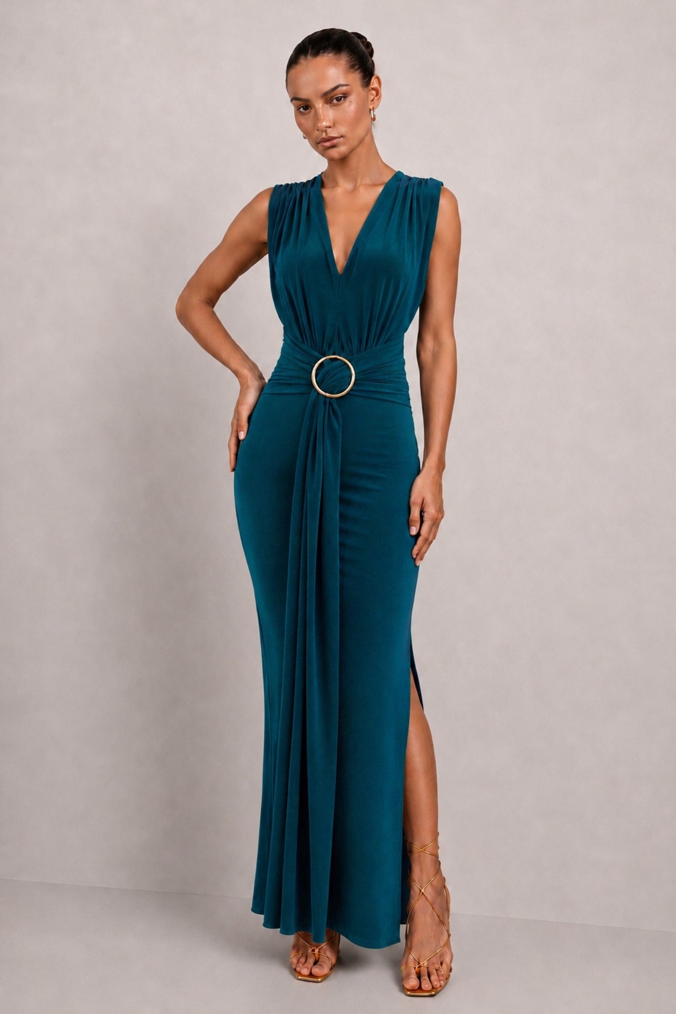 A woman modeling a sleeveless deep V-neck floor-length gown in deep teal with a draped waist and a gold-tone circular belt, standing against a light gray studio backdrop.