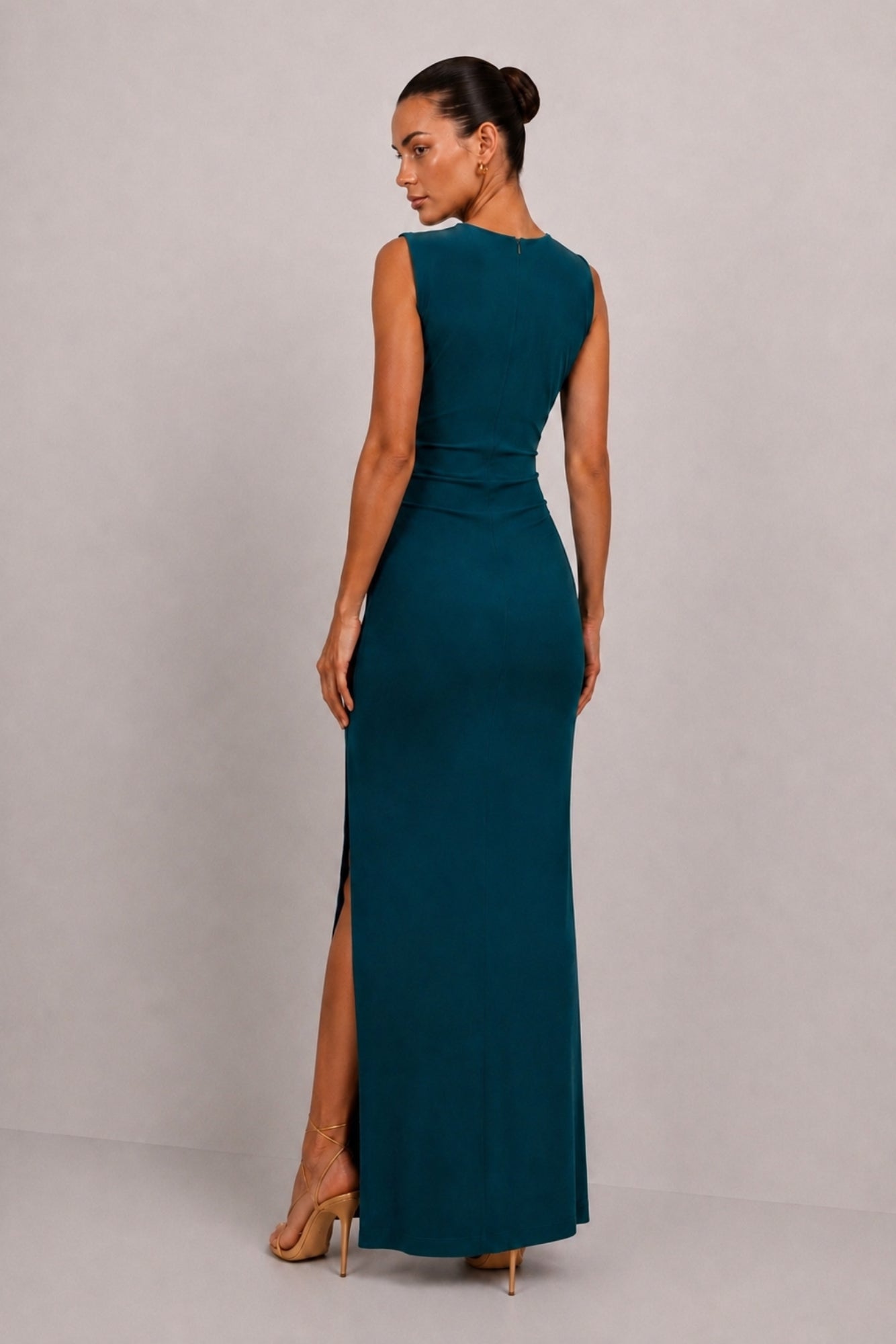 A woman modeling a sleeveless, floor-length teal maxi dress from the back, featuring a high side slit and a fitted silhouette, set against a plain light gray studio background.