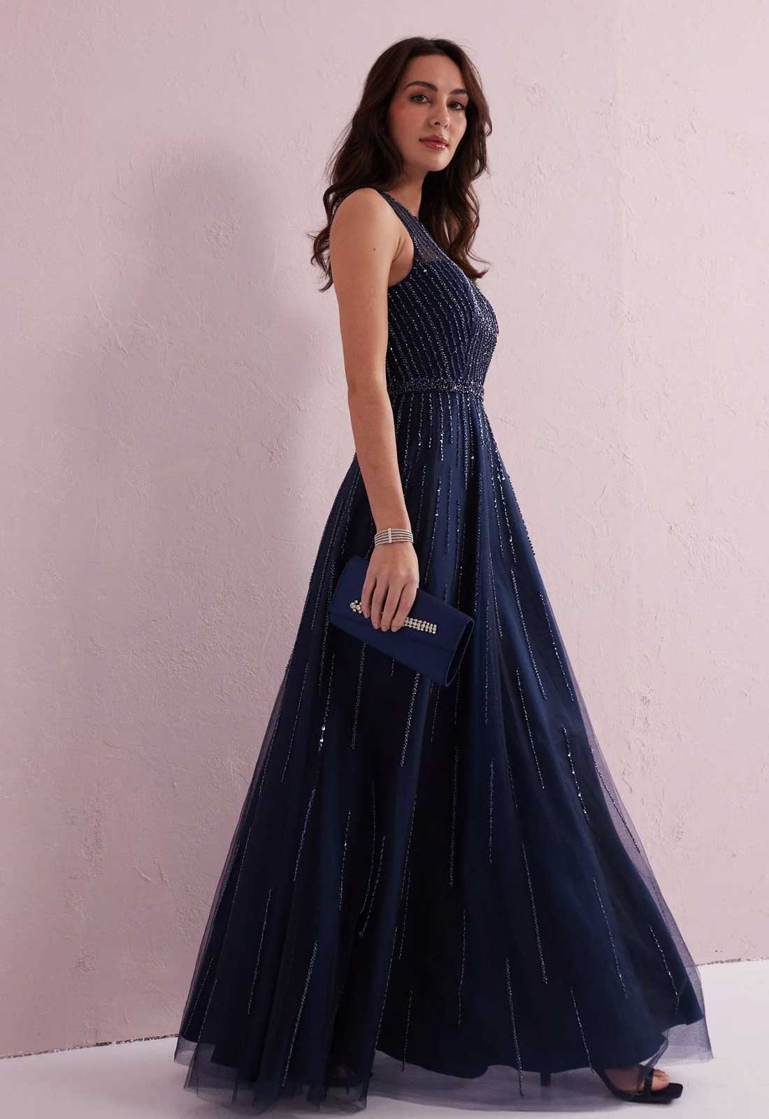 Woman in a navy blue evening gown holding a matching clutch against a light pink background