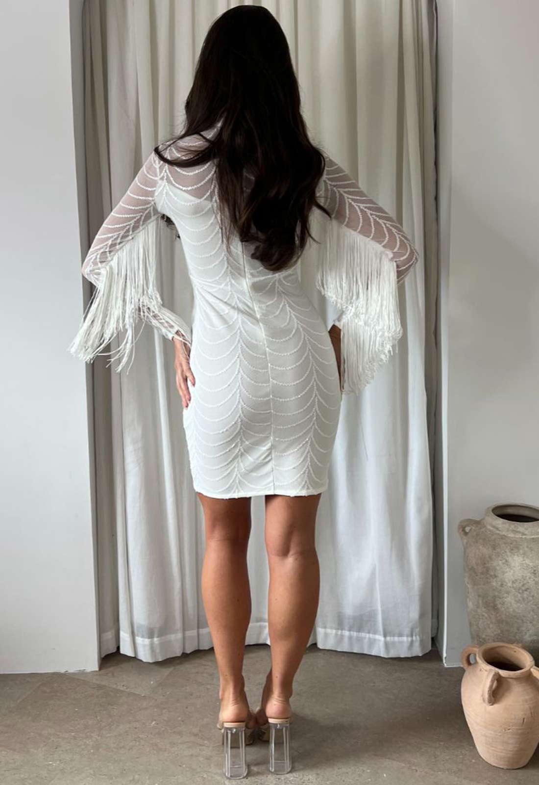 Person wearing a white lace dress with sheer sleeves in a room with curtains and vases.