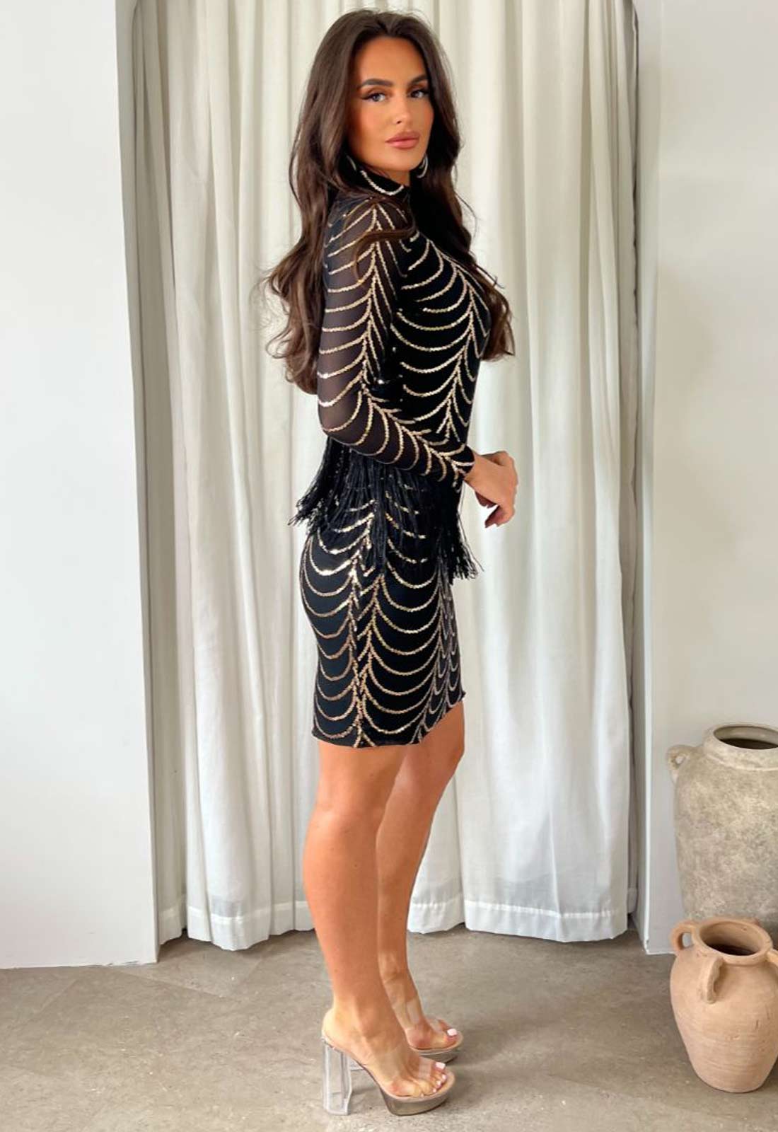 Woman wearing a black and gold dress with sheer sleeves in a room with white curtains.