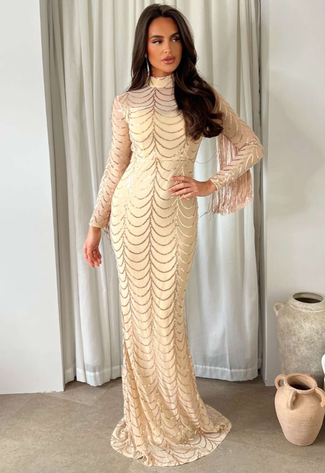 Woman wearing a beige long-sleeve dress with a textured pattern in a room with curtains and a vase.
