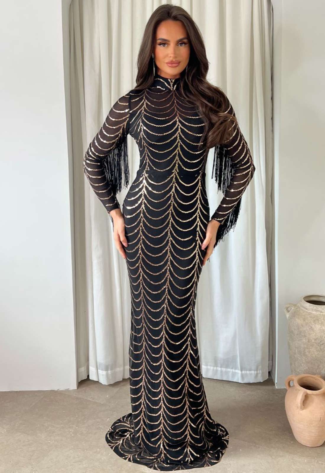 Woman wearing a black and gold patterned dress standing against a white curtain.