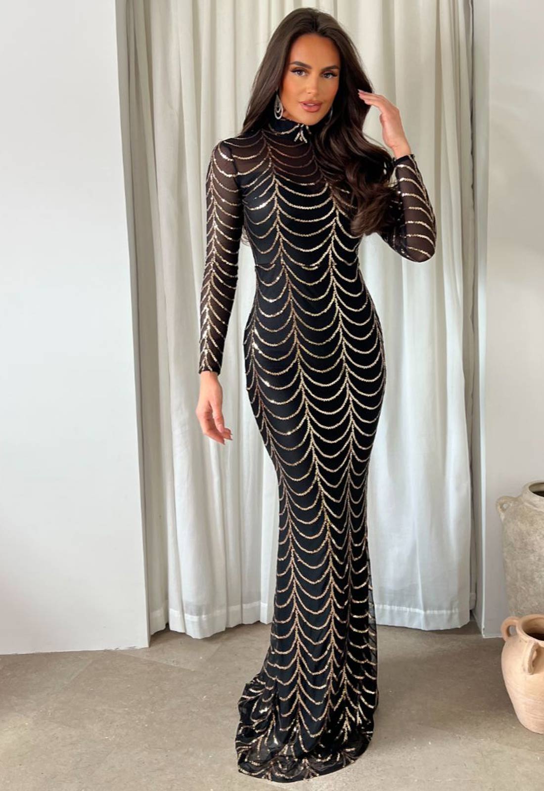 Woman wearing a black and gold patterned evening gown in front of white curtains.