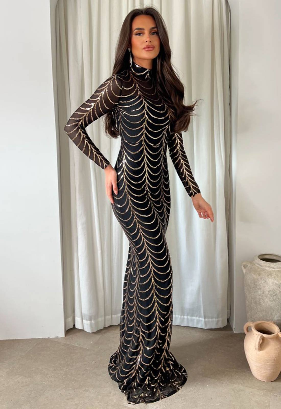 Woman wearing a black and gold patterned dress standing against a white curtain.