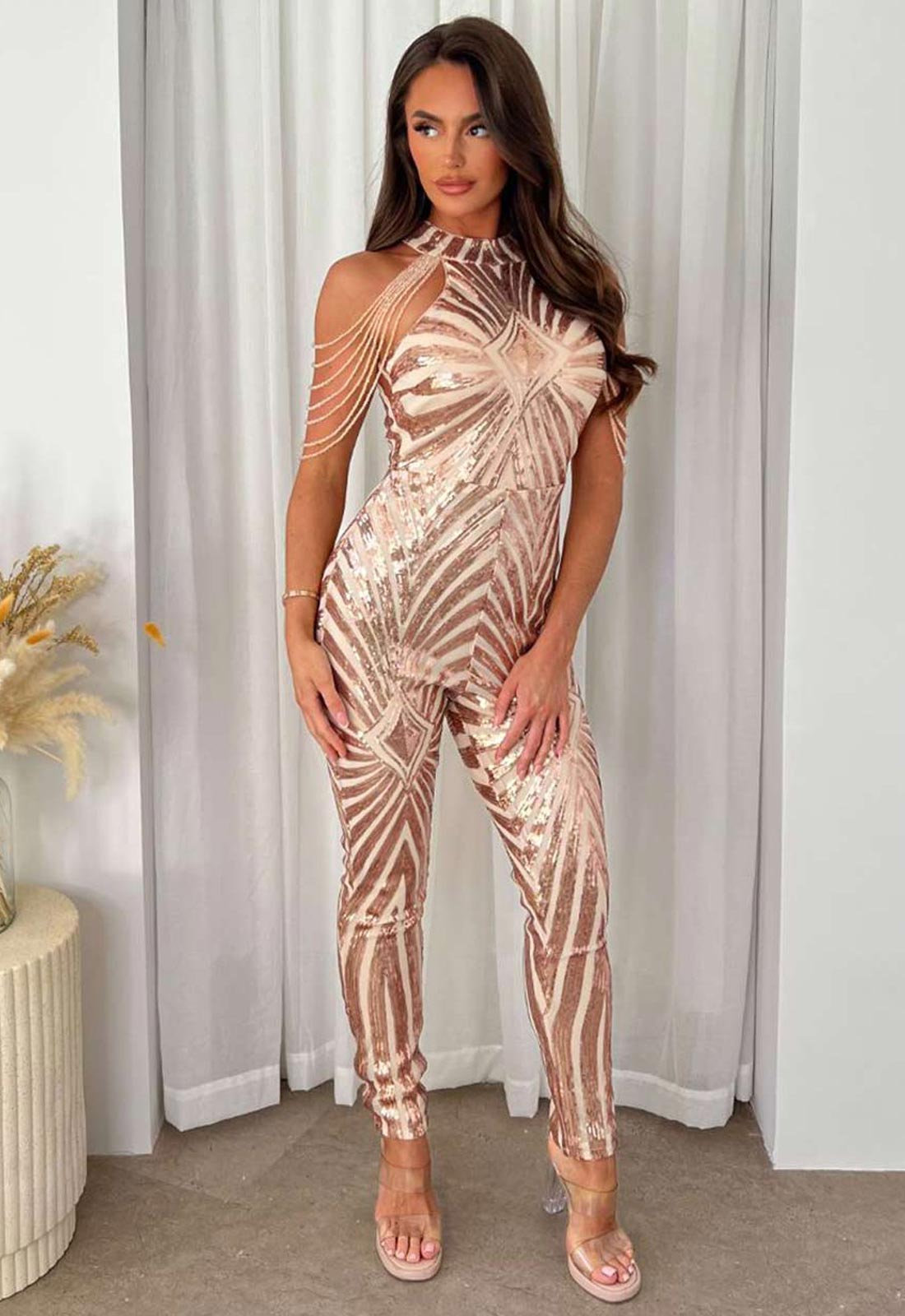 Woman wearing a rose gold sequin jumpsuit standing in front of white curtains.