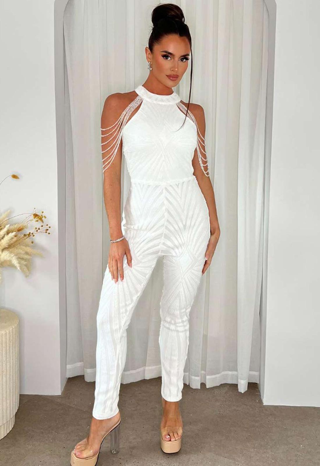 Woman wearing a white jumpsuit with sheer shoulder straps against a neutral background