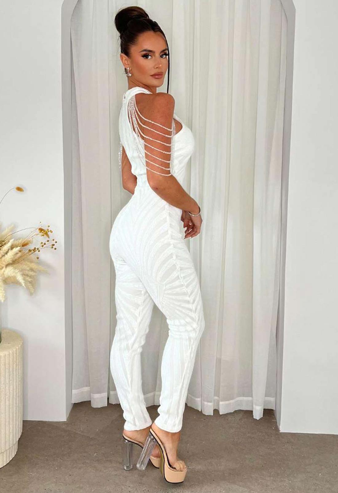 Woman wearing a white open-back jumpsuit standing in front of sheer curtains.