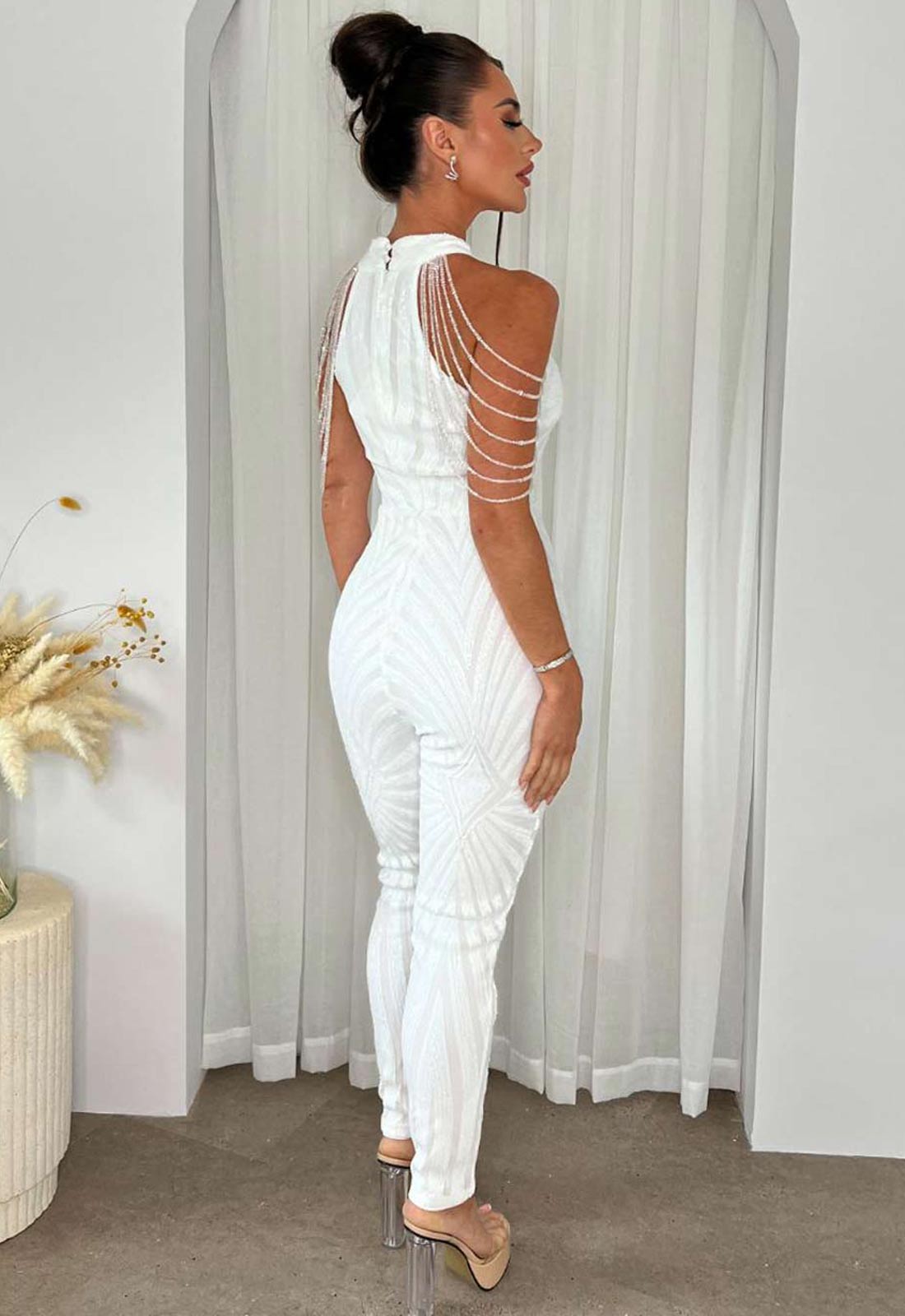 Woman wearing a white jumpsuit with cut-out design in a room with curtains and a plant.