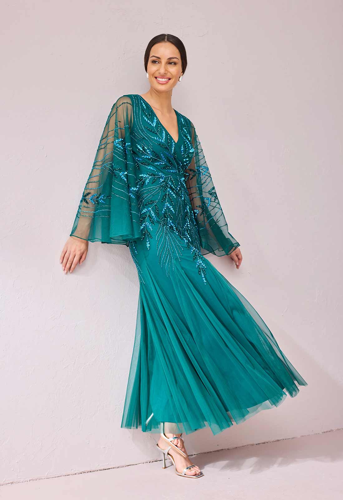 A woman modeling a teal green tulle maxi dress with sheer long sleeves, sequined details, and a flared skirt, posed against a light pink studio wall.