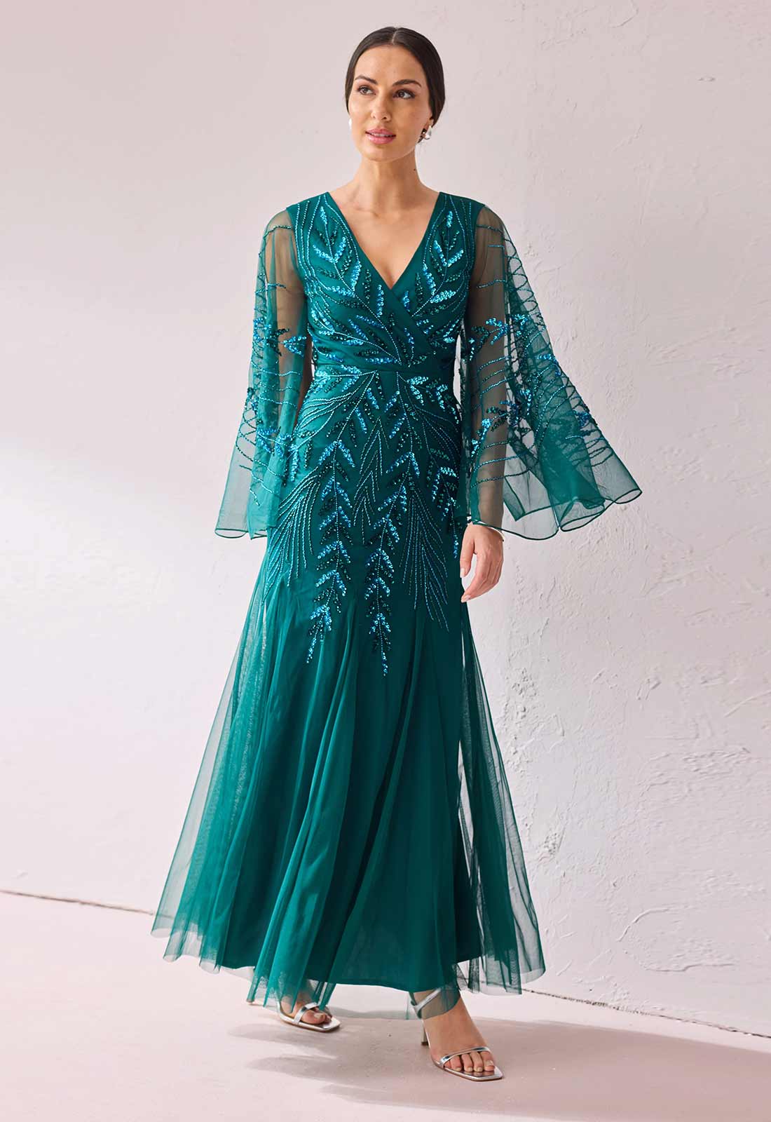 A woman modeling a floor-length teal green evening gown with sheer bell sleeves, a V-neckline, and silver-toned embellishments on the bodice and skirt, standing against a light textured wall.