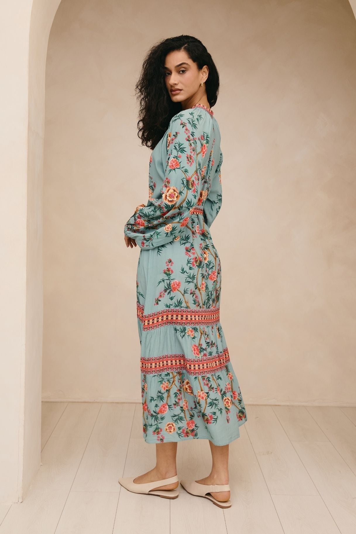 Woman wearing a floral dress standing in a minimalistic room with a beige wall and white floor.