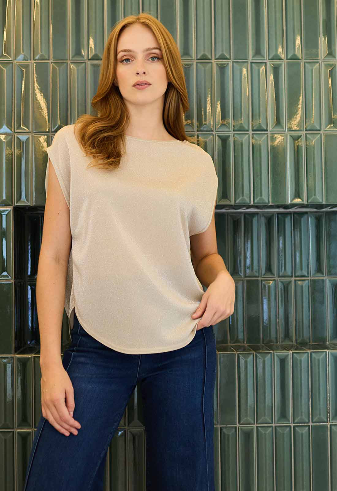 Woman wearing a beige top and blue jeans standing against a green tiled wall.