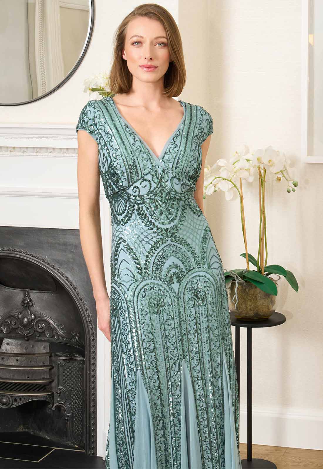 Woman wearing a green patterned dress standing in a room with a fireplace and decorative elements.