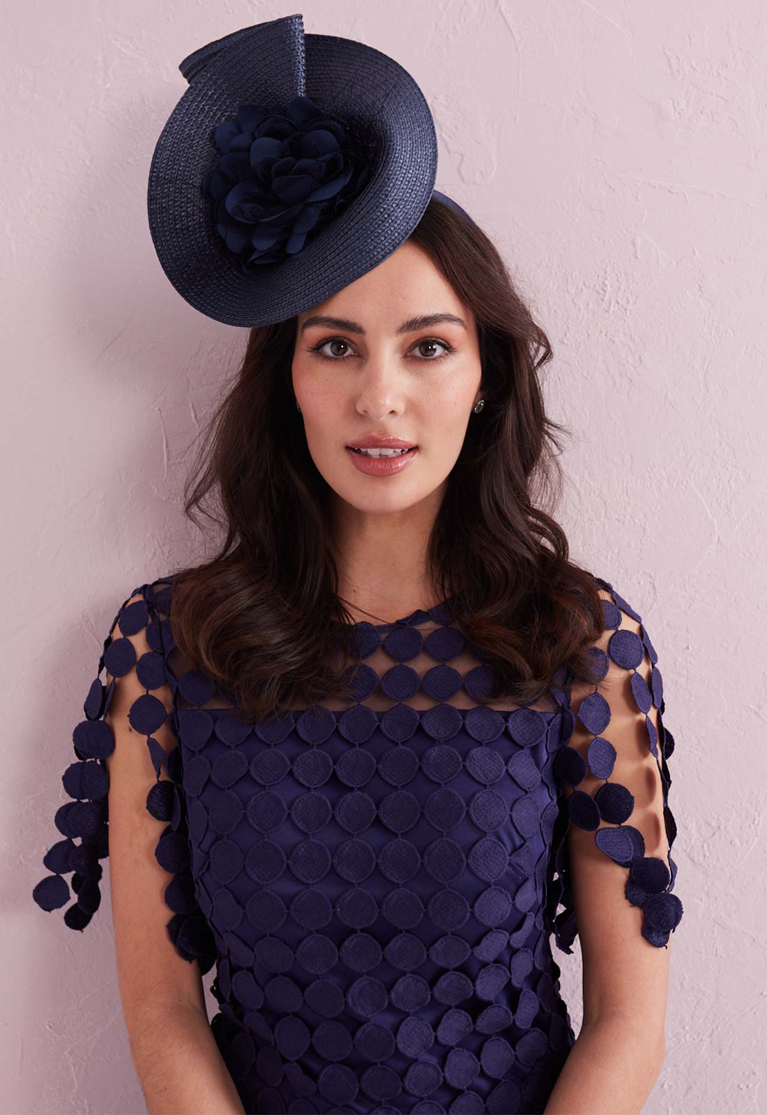 Woman wearing a navy blue dress with floral details and matching hat against a light pink background